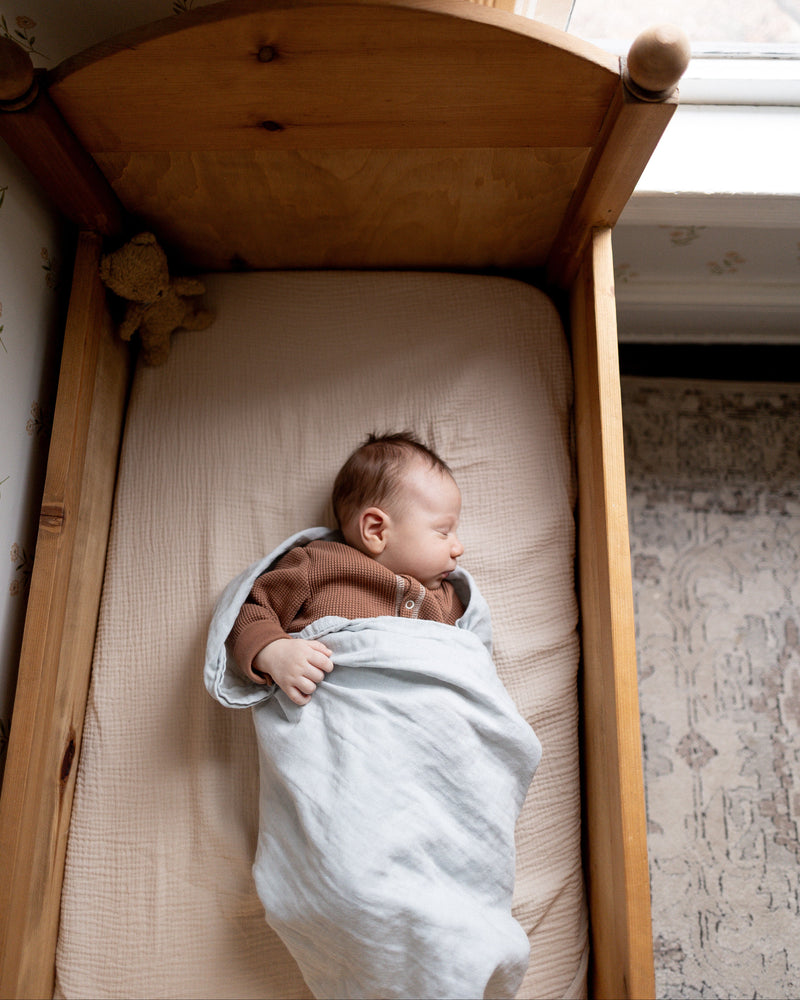 Newborn baby wrapped in a blanket lying in a wooden crib with floral wallpaper in the background.