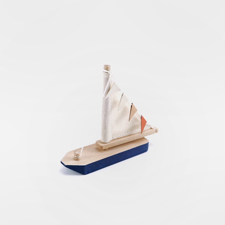 A small wooden toy sailboat with a natural wood hull and mast, white sails, and a dark blue base, photographed on a clean white background.