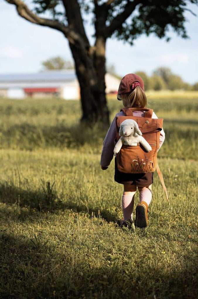 Child walking in a field with a backpack and teddy bear, wearing a red hat.