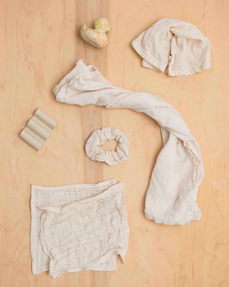 A collection of ivory-colored textiles laid out on a wooden surface, including a large furoshiki, a napkin, a large scrunchie, and two washcloths.